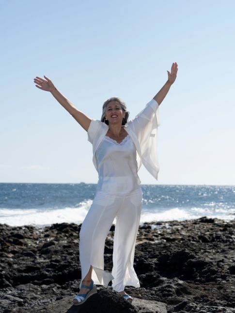 Coaching en Canarias mujer posando playa de piedras, sonriente y con manos levantadas, vestida de blanco